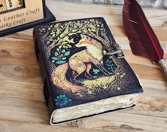 May include: A black leather journal with a colorful illustration of a fox in a forest setting. The journal has a clasp closure and is decorated with intricate details. "Quirky Craft" is printed on the front cover.