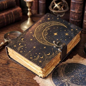 Könnte beinhalten: Ein schwarzes Notizbuch mit festem Einband, das auf der Vorderseite mit einem goldenen Halbmond- und Sternenmuster verziert ist. Das Buch verfügt über einen Metallverschluss und dicke, antik wirkende Papierseiten mit unebenen Kanten.