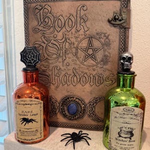 May include: Two glass bottles with cork stoppers and labels. The bottle on the left has a spider web stopper and a label that reads "RARE Spider Legs". The bottle on the right has a skull stopper and a label that reads "Witches' Brew To grow warts". Both bottles are sitting in front of a leather-bound book with the title "Book of Shadows".