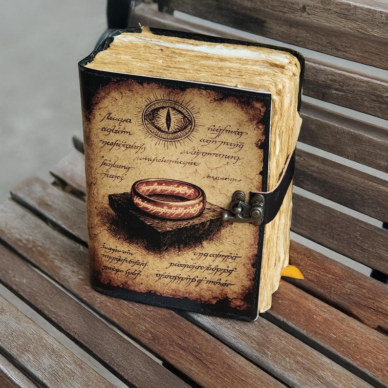 May include: A leather-bound journal with a vintage design. The cover features an illustration of a ring and an eye, with aged paper and a black strap closure. The pages have a yellowed, antique appearance.