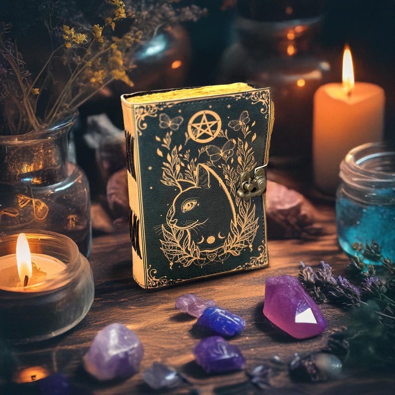 May include: A dark green journal with a cat design, pentagram, and botanical accents. The journal has a clasp closure and gold-colored edges. Amethyst and quartz crystals, candles, and dried flowers surround the journal on a wooden surface.