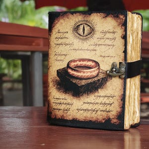 May include: A leather-bound journal with a vintage design. The cover features an illustration of a ring and an eye, with script around the edges. The journal is closed with a black strap and metal clasp. The pages have a yellowed, aged appearance.