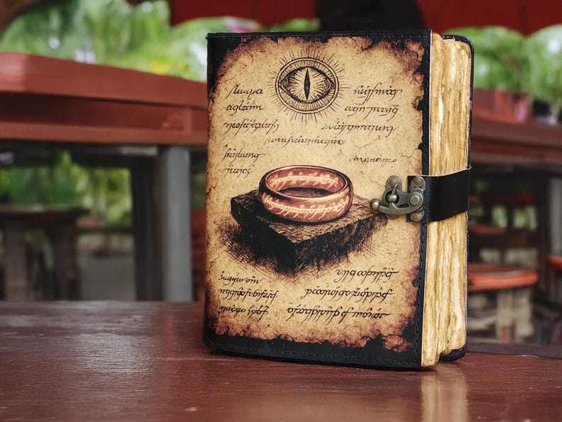 May include: A leather-bound journal with a vintage design. The cover features an illustration of a ring and an eye, with script around the edges. The journal is closed with a black strap and metal clasp. The pages have a yellowed, aged appearance.