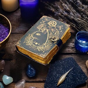 May include: A dark teal leather-bound journal with a cat design and gold accents. The cover features a cat face, botanical elements, and a pentagram. The pages have a golden edge. A dark blue strap secures the journal.