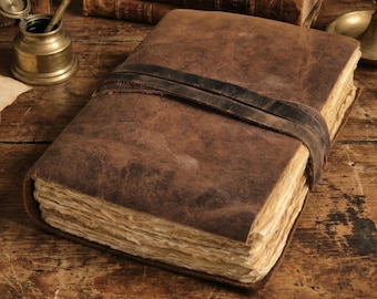 Puede incluir: Un diario de cuero marrón con un cierre de correa de cuero oscuro. El diario tiene páginas envejecidas y texturizadas y está sobre una superficie de madera. Un tintero y una pluma antiguos son visibles.