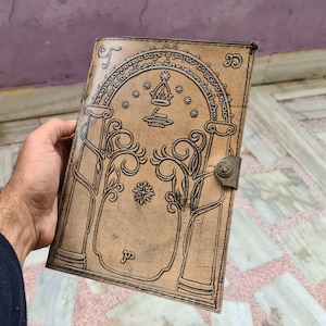 Pode incluir: Um di&aacute;rio de couro castanho claro com um design em relevo com um portal arqueado, &aacute;rvores e estrelas. O di&aacute;rio tem um fecho de metal e uma borda decorativa.