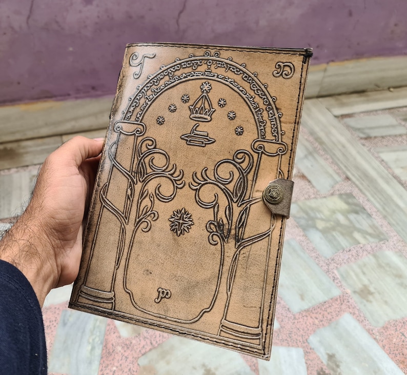 Pode incluir: Um di&aacute;rio de couro castanho claro com um design em relevo com um portal arqueado, &aacute;rvores e estrelas. O di&aacute;rio tem um fecho de metal e uma borda decorativa.