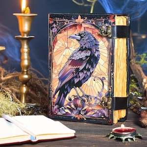 Puede incluir: Un diario decorativo con una ilustración de cuervo. La portada presenta un diseño de estilo vidriera con un cuervo negro, sobre un fondo cálido y dorado. El diario está encuadernado con una correa y un cierre negros, y tiene páginas envejecidas.
