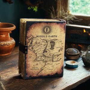 May include: A Middle-earth map journal with a leather strap closure. The cover features a detailed map of Middle-earth with the text "MIDDLE-EARTH" and an eye symbol. The journal has aged, textured pages and is set on a wooden surface.