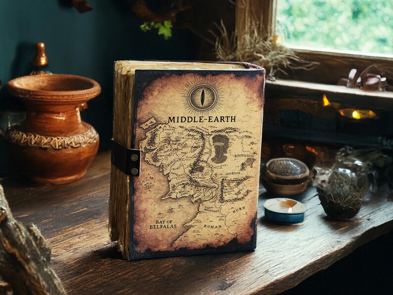 May include: A Middle-earth map journal with a leather strap closure. The cover features a detailed map of Middle-earth with the text "MIDDLE-EARTH" and an eye symbol. The journal has aged, textured pages and is set on a wooden surface.