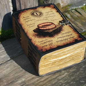 May include: A large, leather-bound journal with a vintage aesthetic. The cover features an illustration of a ring and an eye, with aged parchment-style text. The book is secured with a metal clasp and rests on a wooden surface.