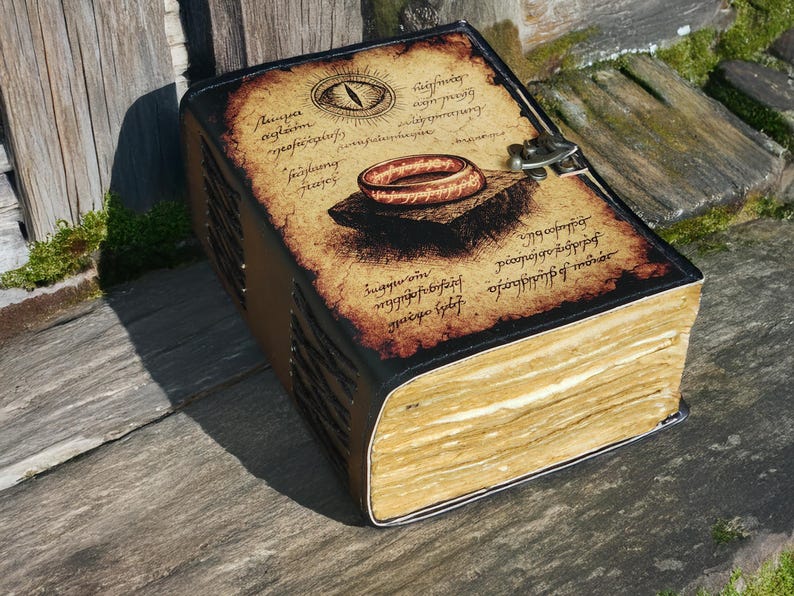 May include: A large, leather-bound journal with a vintage aesthetic. The cover features an illustration of a ring and an eye, with aged parchment-style text. The book is secured with a metal clasp and rests on a wooden surface.