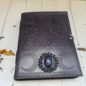 May include: A dark brown leather-bound journal with the words "Book of Shadows" embossed on the cover. The cover also features a pentagram, two crescent moons, and a blue gemstone.