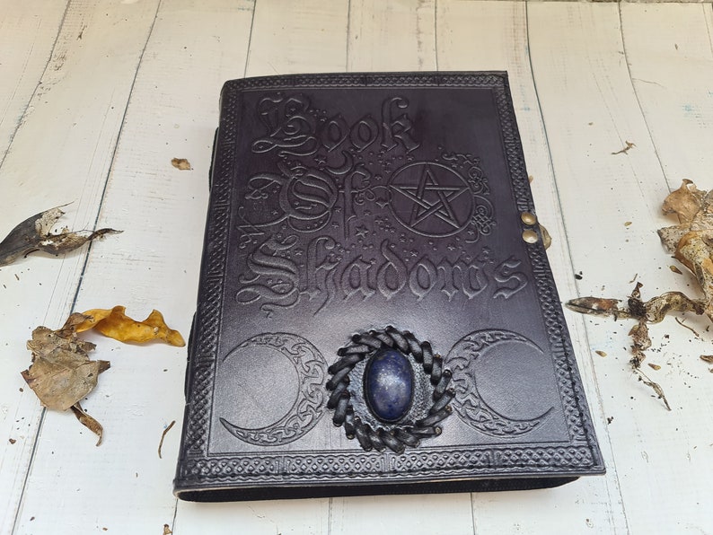 May include: A dark brown leather-bound journal with the words "Book of Shadows" embossed on the cover. The cover also features a pentagram, two crescent moons, and a blue gemstone.