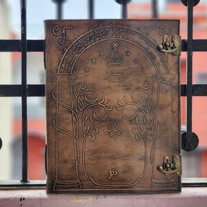 Pode incluir: Um di&aacute;rio de couro castanho com um design em relevo com um arco, &aacute;rvores e estrelas. O di&aacute;rio tem um fecho de fecho e detalhes met&aacute;licos decorativos. A capa tem texto em um padr&atilde;o circular. Um item &uacute;nico, feito &agrave; m&atilde;o.