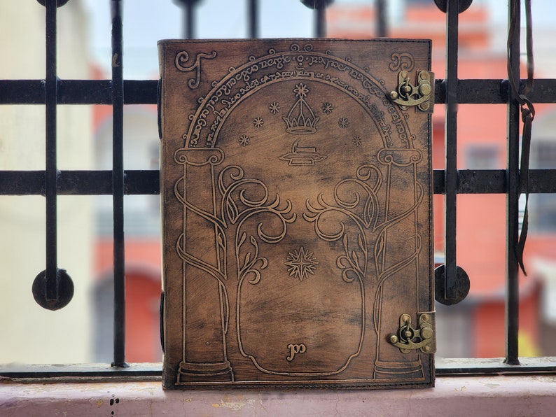 Pode incluir: Um di&aacute;rio de couro castanho com um design em relevo com um arco, &aacute;rvores e estrelas. O di&aacute;rio tem um fecho de fecho e detalhes met&aacute;licos decorativos. A capa tem texto em um padr&atilde;o circular. Um item &uacute;nico, feito &agrave; m&atilde;o.