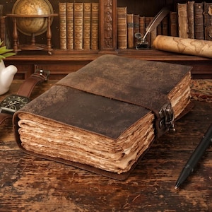 May include: A distressed leather journal with a strap closure. The journal features aged, textured pages and a dark brown cover. A leather strap wraps around the journal, secured by a metal clasp. A watch, pen, and globe are in the background.