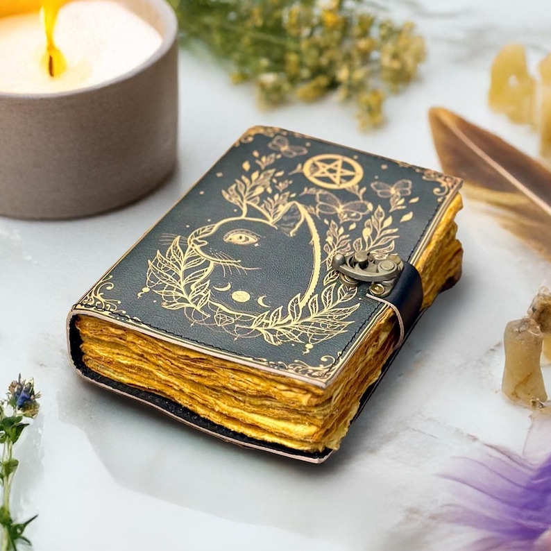 May include: A dark green leather journal with a cat design and gold accents. The cover features a cat silhouette, botanical elements, and a pentagram. The pages have a golden hue, and the journal is secured with a clasp.