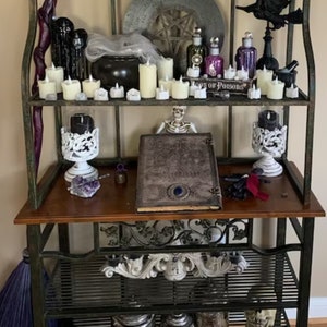 May include: A black metal bakers rack with three shelves. The top shelf has a variety of decorative items, including candles, bottles, and a cauldron. The middle shelf has a book, a skeleton, and two candle holders. The bottom shelf has a decorative metal piece and a few other items.