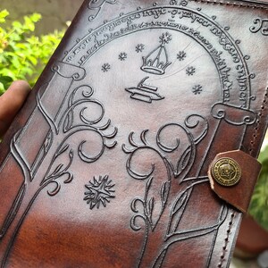 Pode incluir: Um di&aacute;rio de couro castanho com desenhos em relevo. A capa apresenta um desenho arqueado com uma coroa e uma estrela, rodeado por texto. Padr&otilde;es decorativos de &aacute;rvores e estrelas tamb&eacute;m s&atilde;o em relevo. Um bot&atilde;o de lat&atilde;o prende o di&aacute;rio.