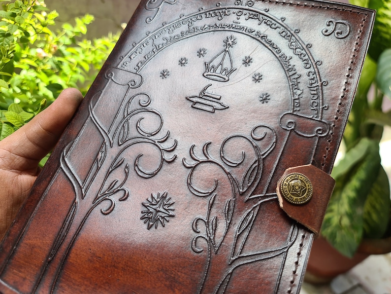 Pode incluir: Um di&aacute;rio de couro castanho com desenhos em relevo. A capa apresenta um desenho arqueado com uma coroa e uma estrela, rodeado por texto. Padr&otilde;es decorativos de &aacute;rvores e estrelas tamb&eacute;m s&atilde;o em relevo. Um bot&atilde;o de lat&atilde;o prende o di&aacute;rio.