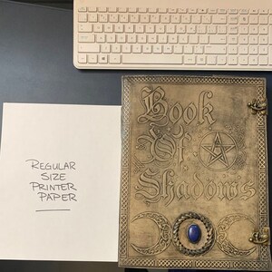 May include: A brown leather-bound journal with the words "Book of Shadows" embossed on the front cover. The cover also features a pentagram, three crescent moons, and a blue gemstone.