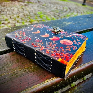 May include: A black leather journal with a floral design and a brass clasp. The cover features a repeating pattern of red and orange moons and stars on a dark background.