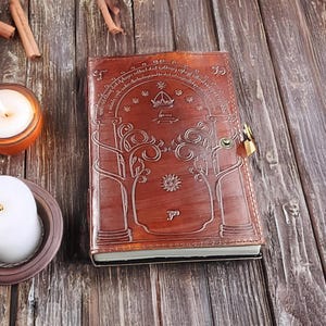 Pode incluir: Um di&aacute;rio de couro castanho com um design em relevo com &aacute;rvores, uma coroa e outros s&iacute;mbolos. O di&aacute;rio tem um fecho com fechadura e chave. A capa &eacute; de uma cor castanha rica e quente, e as p&aacute;ginas s&atilde;o vis&iacute;veis ao longo da borda.