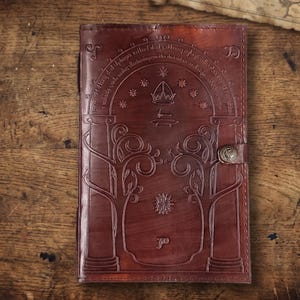 Pode incluir: Um di&aacute;rio de couro castanho com desenhos em relevo, incluindo um arco, &aacute;rvores e estrelas. O di&aacute;rio tem um fecho de fivela e est&aacute; sobre um fundo de madeira. A capa apresenta detalhes intrincados e uma est&eacute;tica vintage.