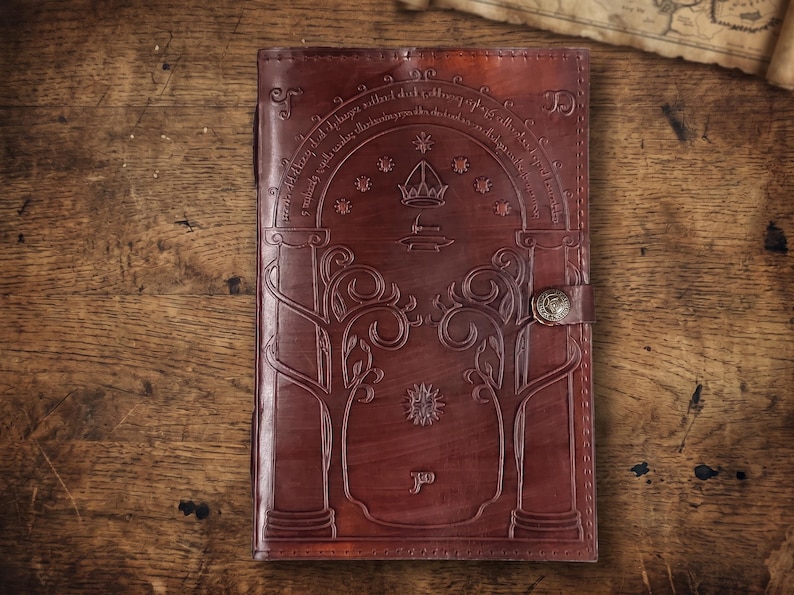 Pode incluir: Um di&aacute;rio de couro castanho com desenhos em relevo, incluindo um arco, &aacute;rvores e estrelas. O di&aacute;rio tem um fecho de fivela e est&aacute; sobre um fundo de madeira. A capa apresenta detalhes intrincados e uma est&eacute;tica vintage.