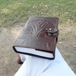 May include: A brown leather journal with a tree of life design and a metal clasp. The journal is closed and sitting on a person's lap.