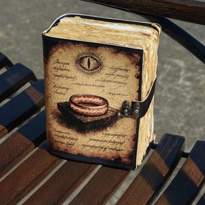 May include: A leather-bound journal with a detailed illustration of a ring and an eye on the cover. The cover has aged paper appearance with script. The journal is closed with a black leather strap and metal clasp. The pages have a yellowed, aged appearance.