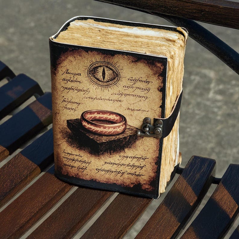 May include: A leather-bound journal with a detailed illustration of a ring and an eye on the cover. The cover has aged paper appearance with script. The journal is closed with a black leather strap and metal clasp. The pages have a yellowed, aged appearance.