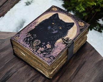 May include: A decorative book with a black wolf illustration on the cover. The cover features a wolf's head with yellow eyes, surrounded by floral and moon designs. The book has a leather strap and clasp.