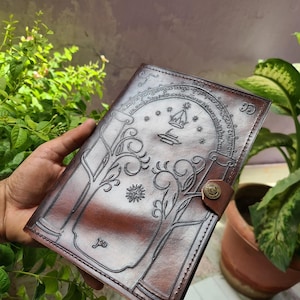 Pode incluir: Um di&aacute;rio de couro castanho com um design em relevo com uma coroa, estrelas e &aacute;rvores estilizadas. O di&aacute;rio tem um fecho de bot&atilde;o e detalhes intrincados. A capa &eacute; de cor castanha escura.