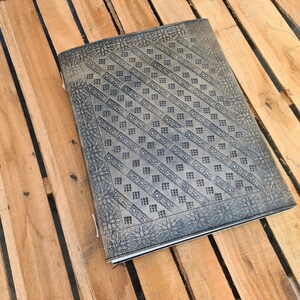 May include: A brown leather journal with a decorative embossed pattern. The pattern features a repeating geometric design with a floral border.
