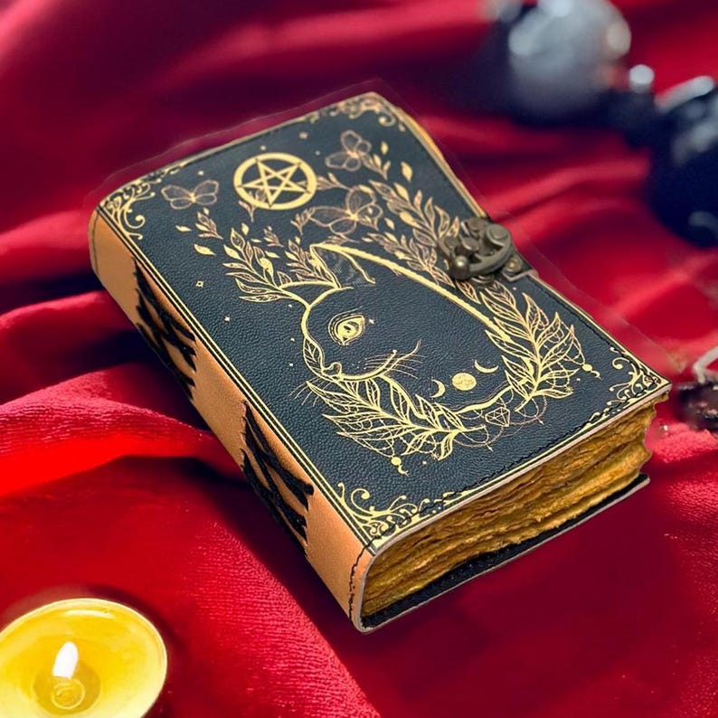 May include: A dark blue leather-bound journal with a gold rabbit design, a pentagram, and botanical accents. The journal has a clasp closure and aged-looking pages. The edges are tan, and the journal is on a red surface.