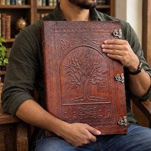 May include: A large, rectangular, dark brown leather-bound book featuring an embossed tree of life design on the front cover. The cover includes intricate border patterns, decorative corner motifs, and three antique-style metal latches on the side.