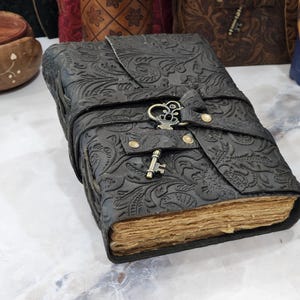 Könnte beinhalten: Ein schwarzes Lederjournal mit einem geprägten Blumenmuster und einem Schlüsselsverschluss. Das Journal hat einen rustikalen, Vintage-Look und ist perfekt zum Aufschreiben von Gedanken und Ideen.