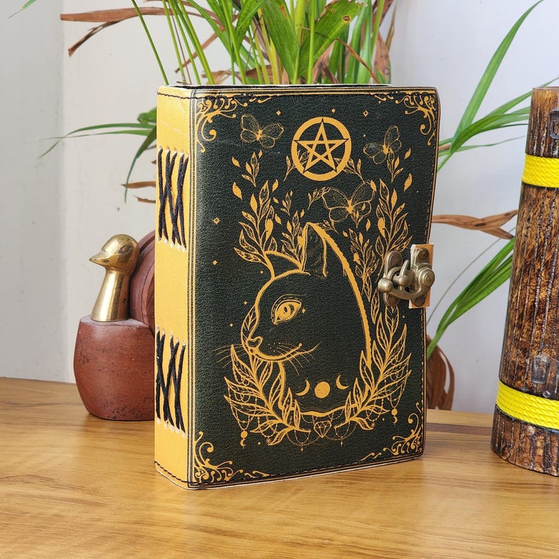 May include: A dark green journal with a gold cat illustration, pentagram, and butterfly accents. The spine is yellow with black stitching. It has a metal clasp closure. The journal is on a wooden surface.