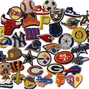May include: A collection of assorted rubber shoe charms featuring various sports team logos, basketball icons, football helmets, baseballs, sneakers, and player silhouettes in a wide array of colors including red, blue, yellow, black, and white.