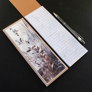 May include: A rectangular notepad featuring a watercolour illustration of plants and grasses in brown, grey, and white. The notepad is open, showing lined paper, with a black pen resting alongside. The notepad has a brown cardboard backing.