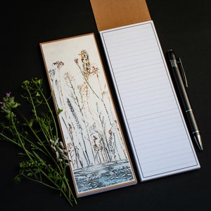 May include: A notepad with a brown cover and a white lined interior. The notepad has a floral design on the front cover with a watercolour effect. The design features brown, orange, and white flowers and stems.