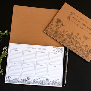 May include: A weekly planner with a floral design. The planner has a brown cover and a white interior with spaces for each day of the week. The text "What's on this week" is printed on the cover and the interior.