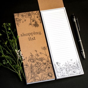 May include: A brown paper shopping list notepad with a floral design and the text "shopping list" printed on the front. The notepad has white lined pages and is accompanied by a black pen.