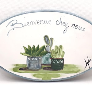 Puede incluir: Un letrero ovalado de cerámica blanca con borde azul, que presenta el texto "Bienvenue chez nous" en escritura cursiva. El letrero está decorado con una ilustración pintada a mano de tres suculentas en macetas en tonos verdes y azules.