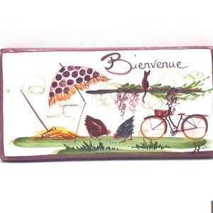 Può includere: Una piastrella di ceramica rettangolare con una scena dipinta di una bicicletta, un gatto, due galline, un rastrello e un ombrello. La piastrella ha un bordo rosa e le parole "Bienvenue" sono dipinte in nero.