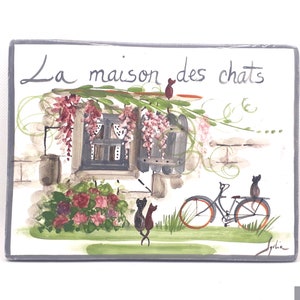 May include: Rectangular ceramic plaque with a hand-painted scene of a house with a window, flowers, and cats. The text "La maison des chats" is written across the top. A bicycle is parked nearby.
