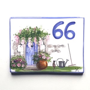 Pode incluir: Placa de número de casa de cerâmica pintada à mão com uma porta azul, flores cor-de-rosa e um gato. O número 66 está pintado de azul na placa.