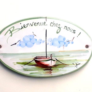 May include: Oval ceramic sign with a green border and a painted scene of a sailboat on a body of water with blue clouds in the sky. The sign reads "Bienvenue chez nous!" in French.
